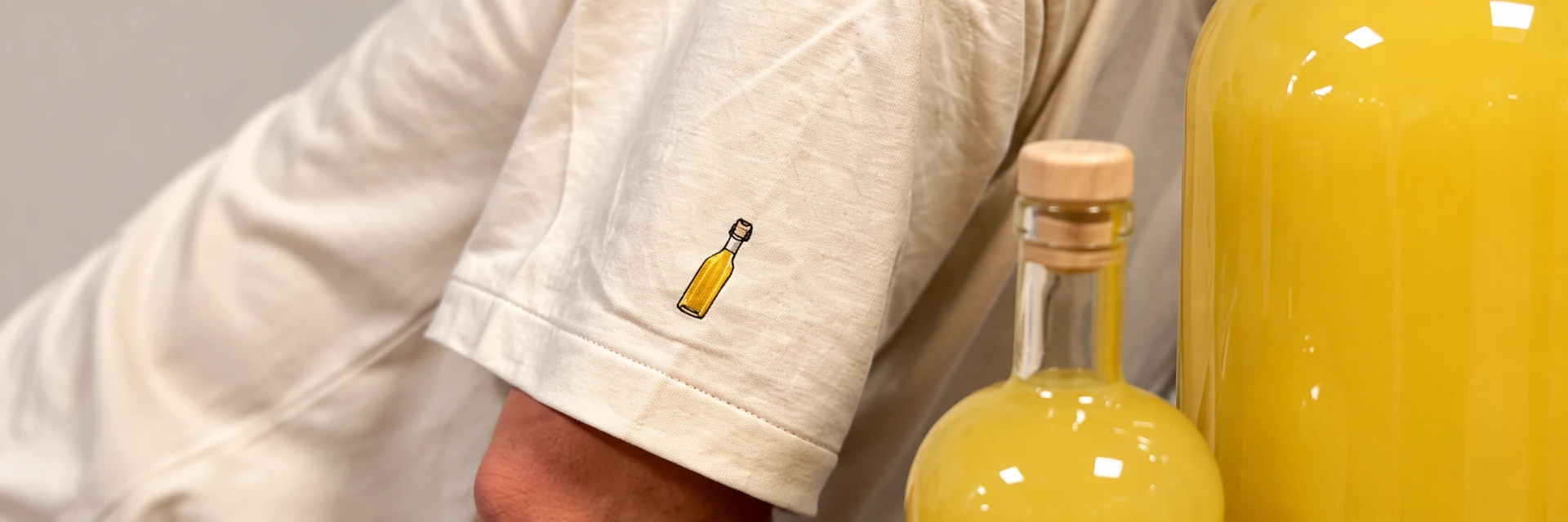 Mouw van een natur tenue met klein geborduurd flesje limoncello, ernaast flessen limoncello